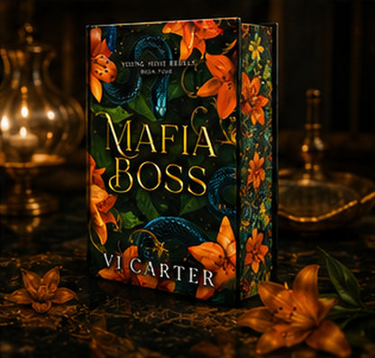 A boxed book or book set titled "Mafia Boss" by Vi Carter, shown standing upright on a dark surface with ornate brass lamps and flowers around it. The cover art features dark green and black tones with orange flowers, blue snakes, and gold lettering.