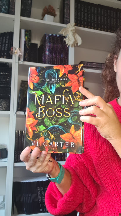 A person in a bright pink sweater holds up a book titled 'Mafia Boss' by Vi Carter, showing the colorful cover with orange flowers and green snakes against a background of bookshelves.